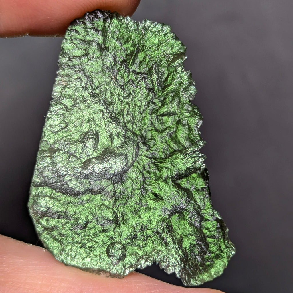 Natural Moldavite from the Olešnice locality weighing 4.69 g with flat elongated shape, sharply sculpted grooves, hammered texture effect and naturally rounded edge in hand - front side, backlight.