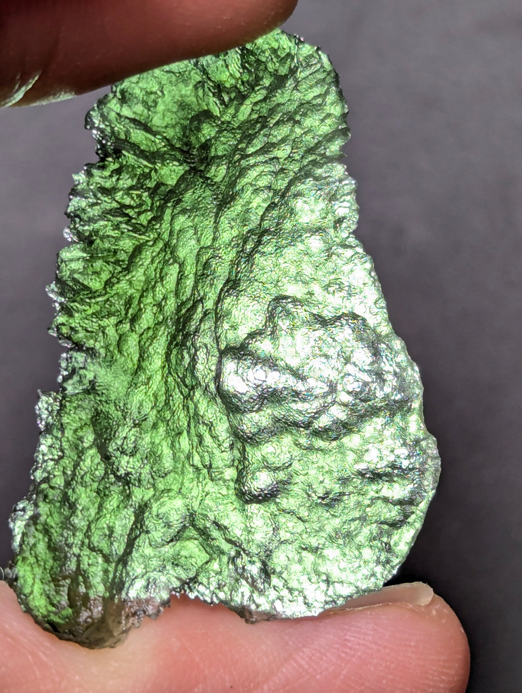 Natural Moldavite from the Olešnice locality weighing 4.69 g with flat elongated shape, sharply sculpted grooves, hammered texture effect and naturally rounded edge in hand - back side, backlight.