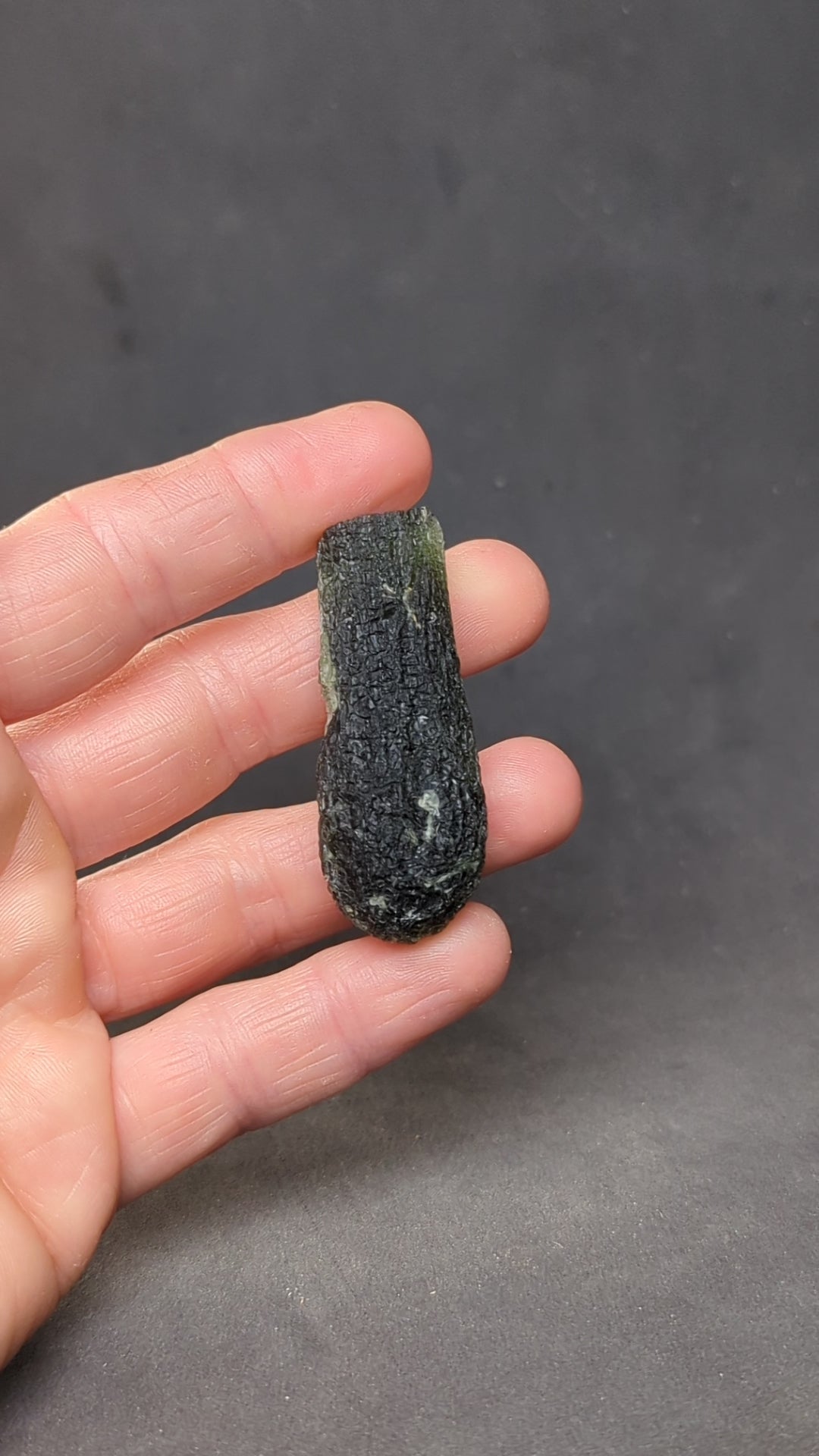 Natural Moldavite from the Jankov locality in hand, front view with elongated drop-like shape, tree bark sculpture, dark olive green translucency and authentic natural relief. Weight 15.62 g - video.