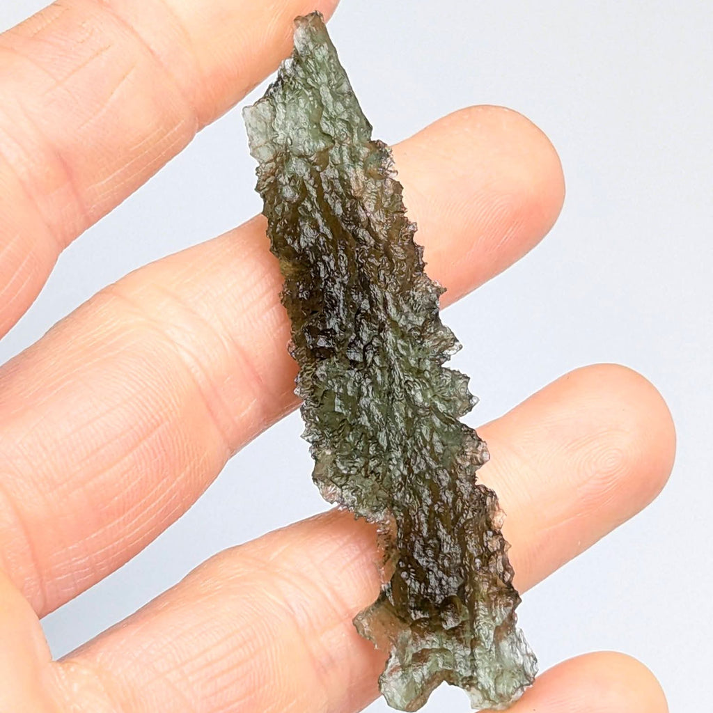 Natural collector moldavite from Dobrkovská Lhotka – Bažina 5.19 g with deep sculptation, elongated shape, and dark olive green color, photographed between fingers - front view.