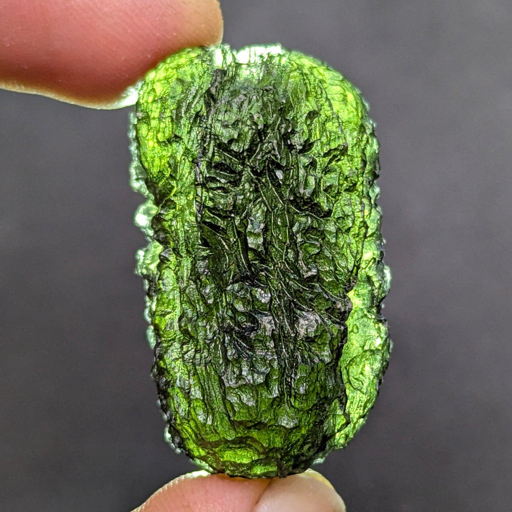 Natural Moldavite from the Olešnice locality with dramatic sculpture, deep central groove, naturally opened bubbles and vivid green translucency under illumination in hand - back side, backlight.