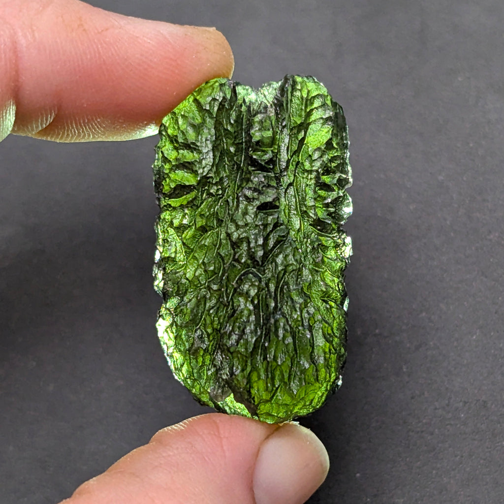 Natural Moldavite from the Olešnice locality with dramatic sculpture, deep central groove, naturally opened bubbles and vivid green translucency under illumination in hand - front view, backlight.