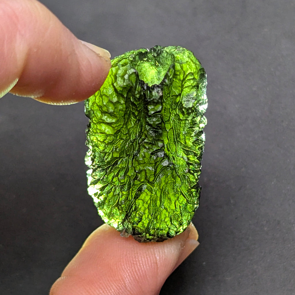 Natural Moldavite from the Olešnice locality with dramatic sculpture, deep central groove, naturally opened bubbles and vivid green translucency under illumination in hand.