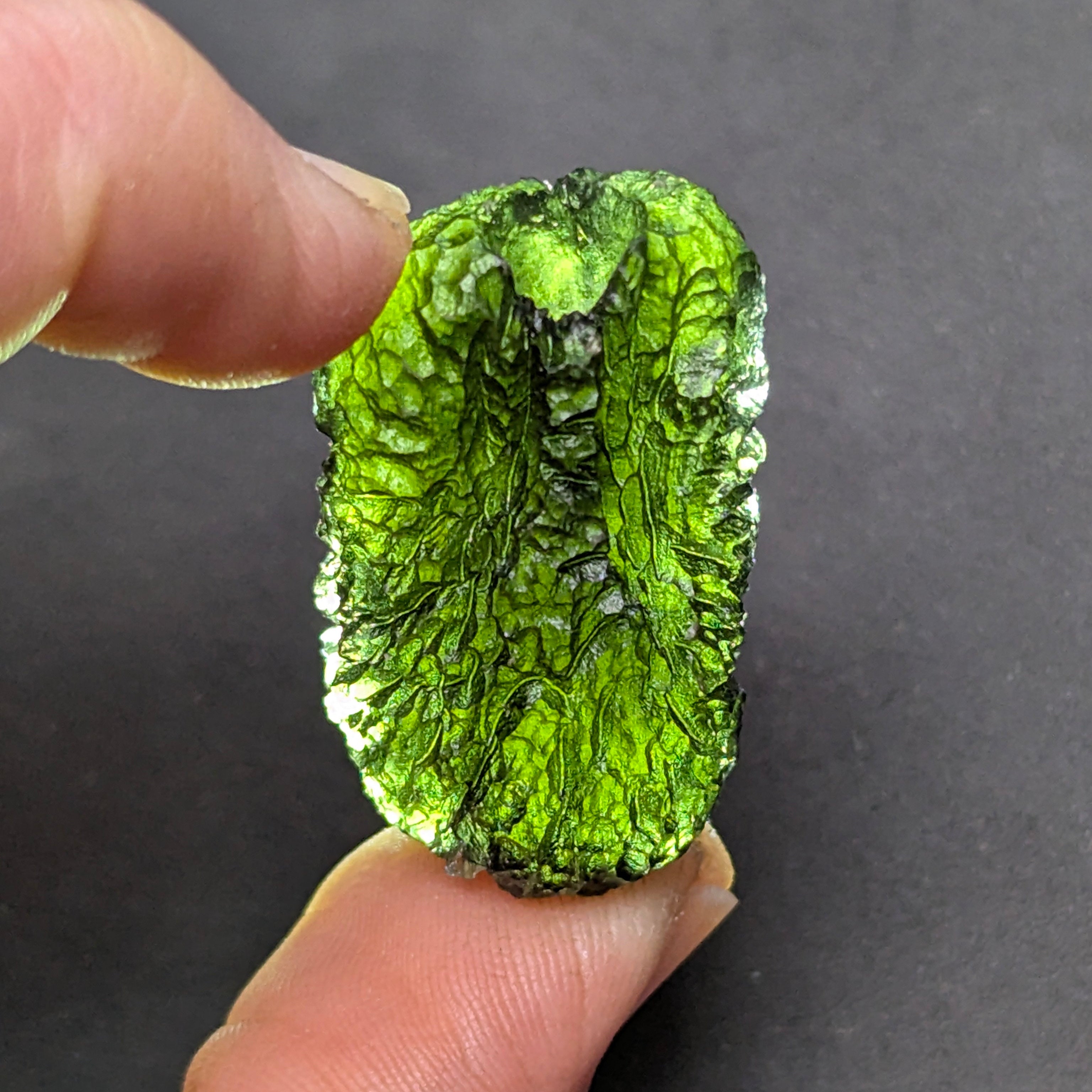 Natural Moldavite from the Olešnice locality with dramatic sculpture, deep central groove, naturally opened bubbles and vivid green translucency under illumination in hand.