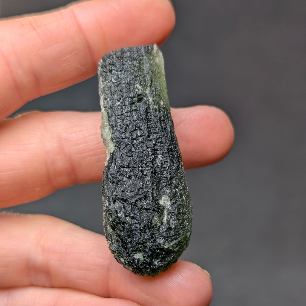 Natural Moldavite from the Jankov locality in hand, front view with elongated drop-like shape, tree bark sculpture, dark olive green translucency and authentic natural relief. Weight 15.62 g - front view.