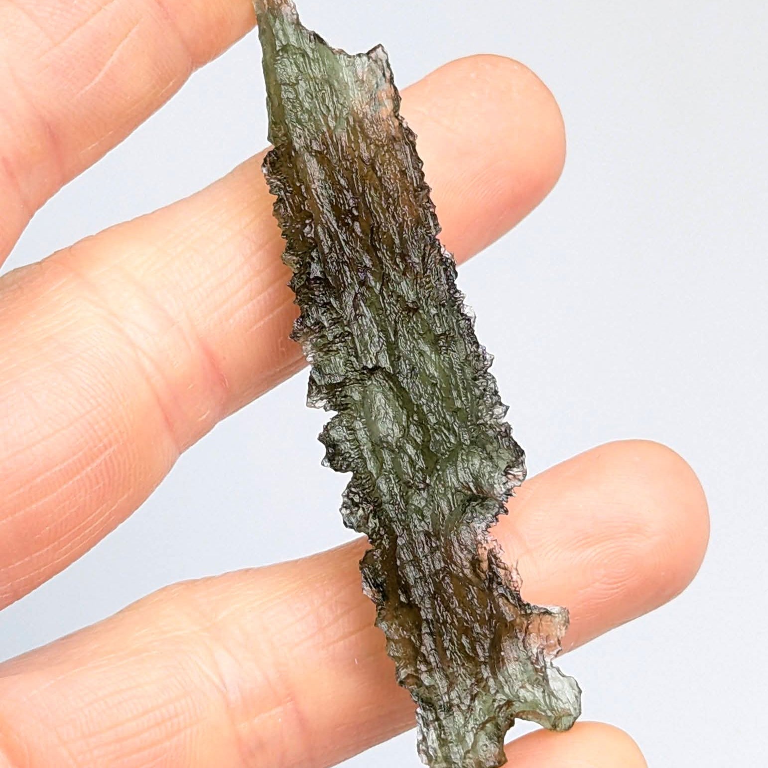 Natural collector moldavite from Dobrkovská Lhotka – Bažina 5.19 g with deep sculptation, elongated shape, and dark olive green color, photographed between fingers - back side.