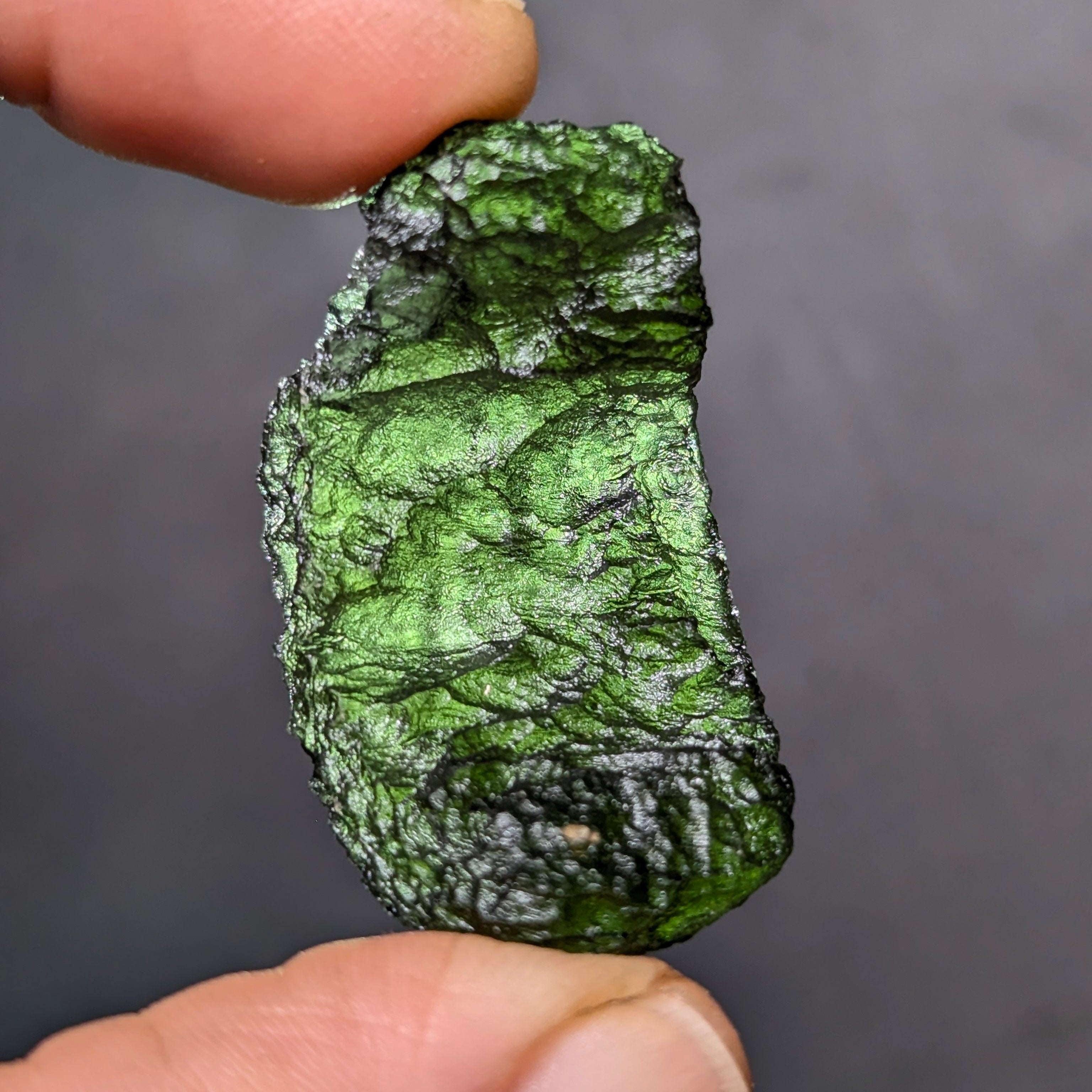 Natural Moldavite from the Olešnice locality weighing 8.86 g with hollow shape, open bubble formation, matte to velvety surface, subtle glossy areas and olive green color in hand - back side, backlight.