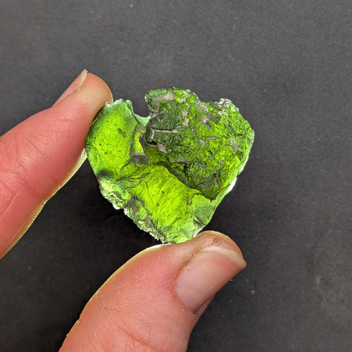 Heart-shaped natural raw moldavite with newly formed sculpture after natural separation, velvety matte surface and fine pyramidal micro-sculpture in hand - front view, backlight.