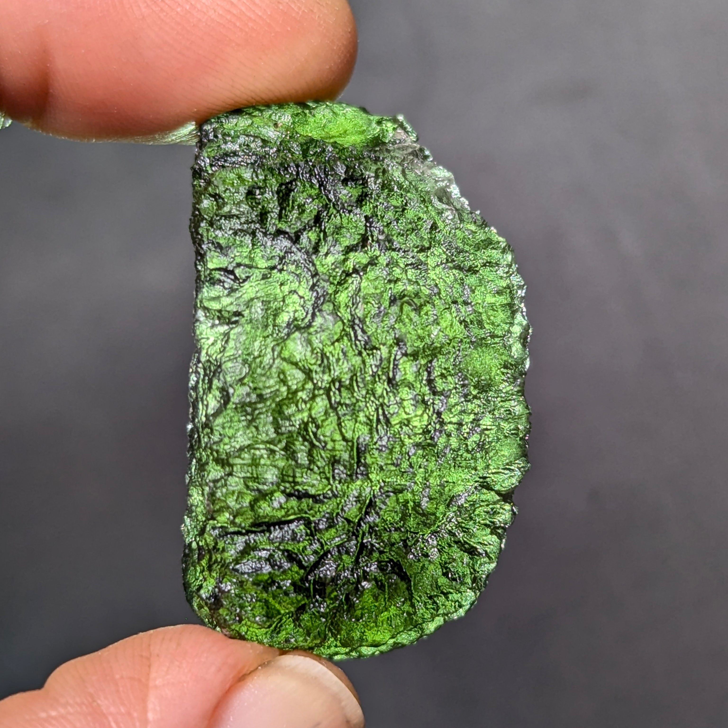 Natural Moldavite from the Olešnice locality weighing 8.86 g with hollow shape, open bubble formation, matte to velvety surface, subtle glossy areas and olive green color in hand - front view, backlight.