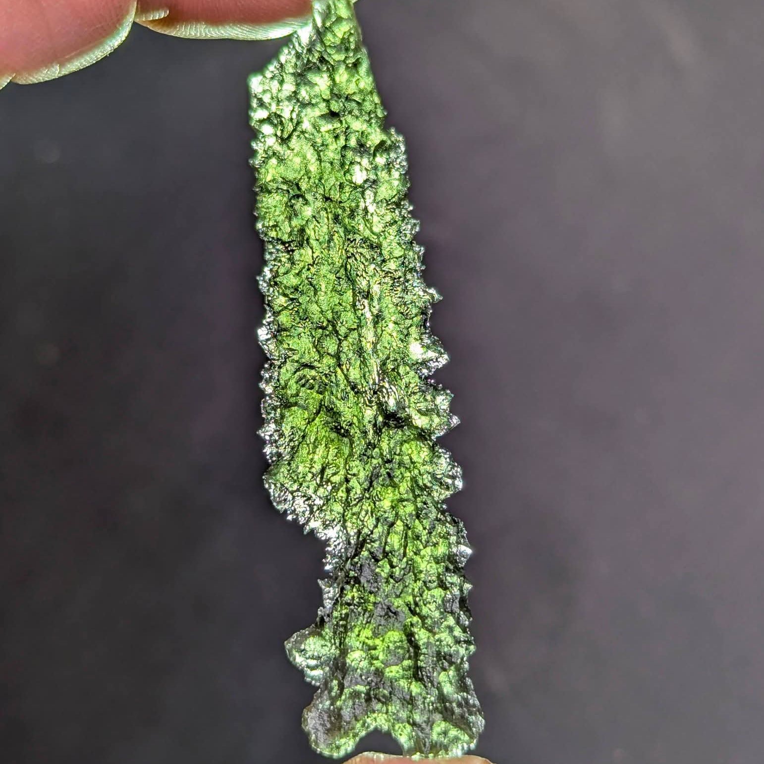 Natural collector moldavite from Dobrkovská Lhotka – Bažina 5.19 g with deep sculptation, elongated shape, and dark olive green color, photographed between fingers - front view, backlight.
